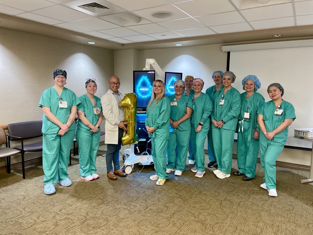 A group of healthcare workers in green scrubs and one in a white coat stand together indoors, smiling. One person holds a gold balloon shaped like the number 1; medical equipment and a screen are in the background.