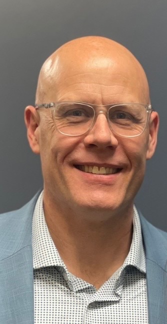 A smiling, bald man wearing clear glasses, a light blue suit jacket, and a white patterned shirt stands in front of a plain gray background.