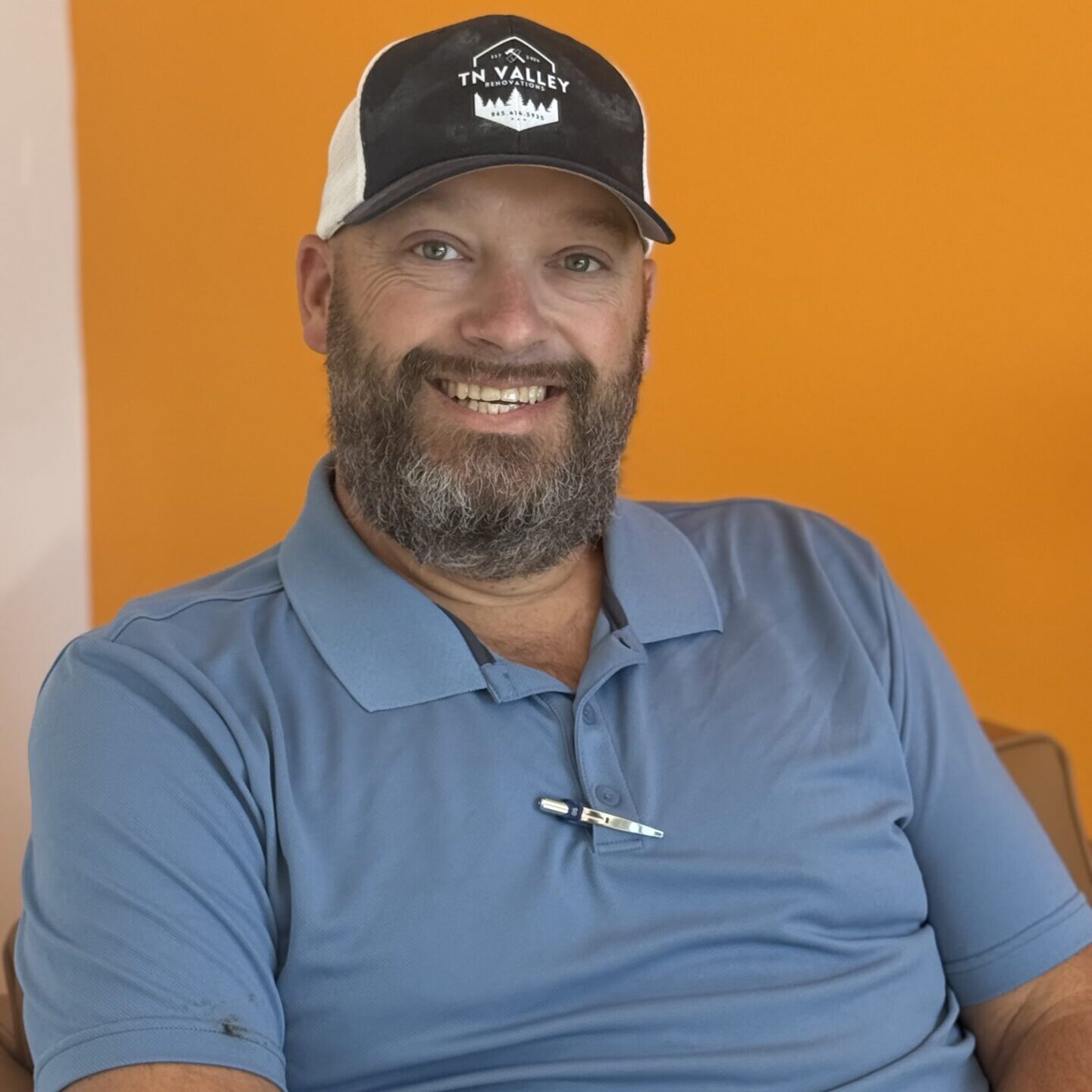 A smiling man with a beard wears a blue polo shirt and a black-and-white cap while sitting against a bright orange background. A pen is clipped to his shirt.