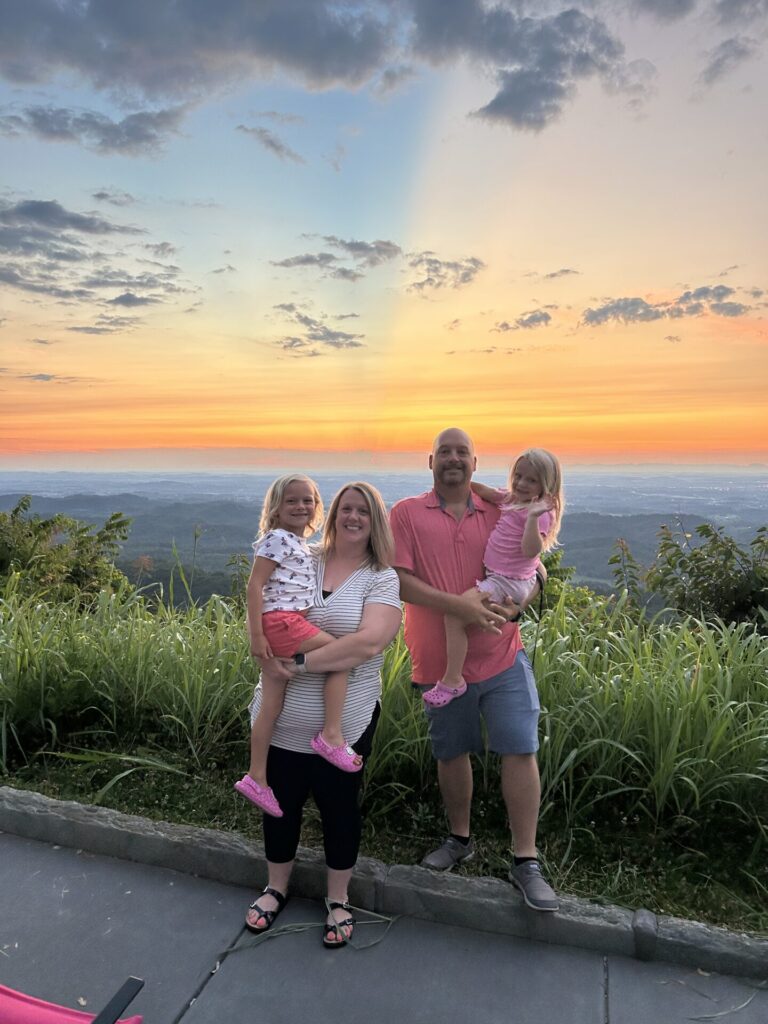 A smiling family of four poses outdoors at sunset, with lush green grass and a scenic view behind them. Two adults each hold a young child, and the sky is colorful with clouds and orange, yellow, and blue hues.