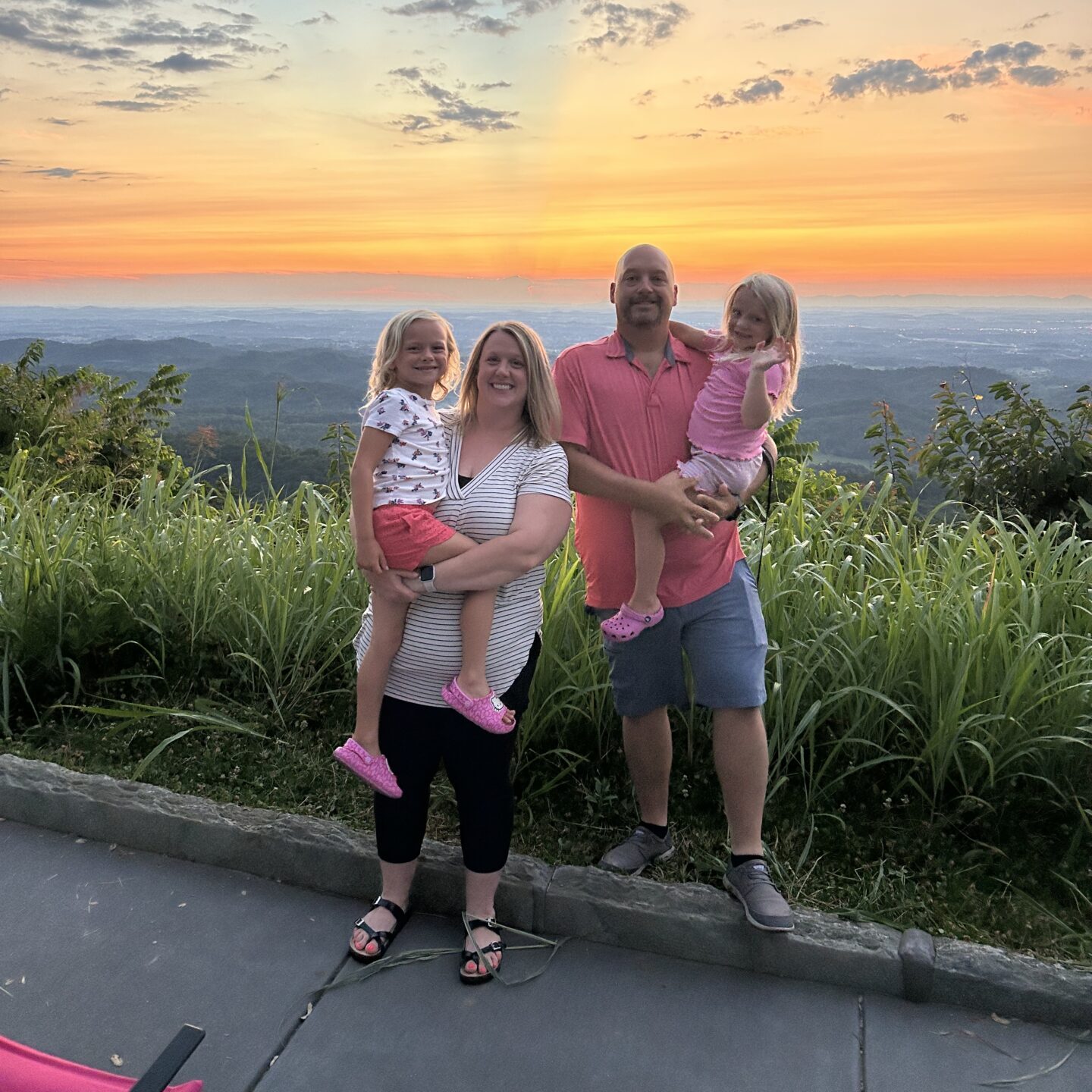 A smiling family of four poses outdoors at sunset, with lush green grass and a scenic view behind them. Two adults each hold a young child, and the sky is colorful with clouds and orange, yellow, and blue hues.