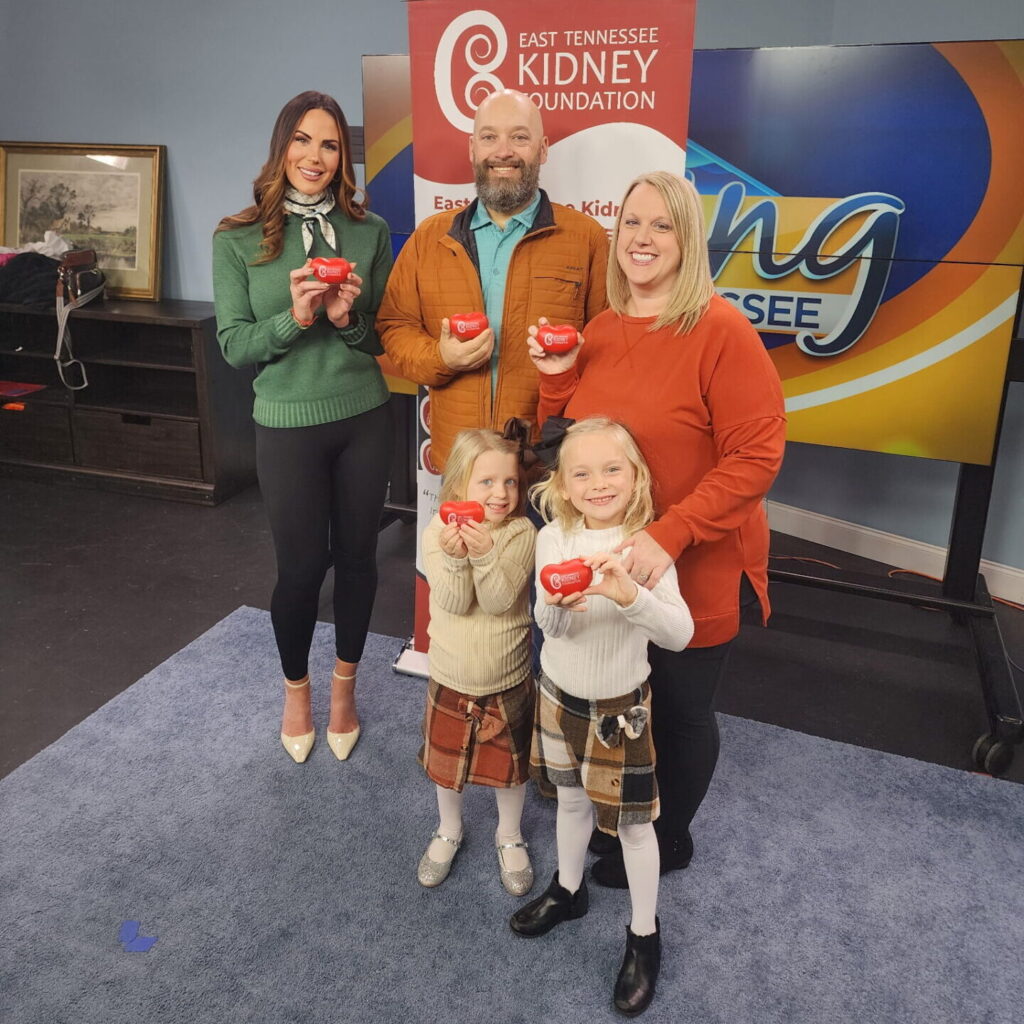 Five people, including two smiling young girls, stand indoors holding red heart-shaped objects. A banner for the East Tennessee Kidney Foundation and a Living East Tennessee sign are visible in the background.