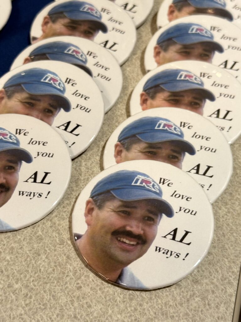 Several round buttons featuring a smiling man wearing a blue cap, with the text We love you ALways! printed beside his photo, are laid out on a table.
