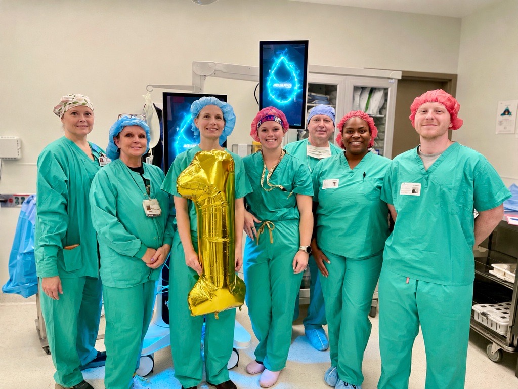 Seven medical professionals in scrubs and colorful caps stand together in a hospital room, smiling as they celebrate one year of Aquablation therapy in Knoxville. One holds a gold balloon shaped like the number 1, with medical equipment visible behind them.
