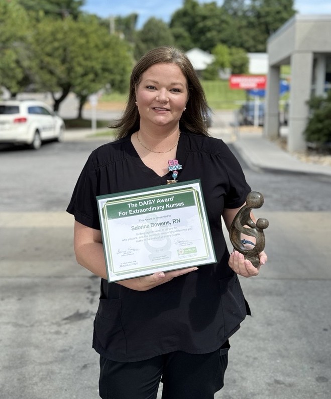 A woman in black scrubs stands outdoors holding a framed certificate titled The DAISY Award For Extraordinary Nurses and a small award statue. She is smiling, with trees and buildings in the background.