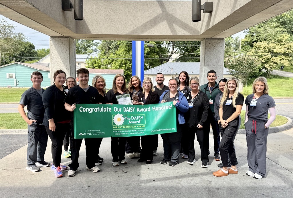 A group of medical staff pose outside under a canopy, smiling and holding a green banner that reads Congratulations Our DAISY Award Honoree! The group appears to be celebrating a nurse’s recognition.