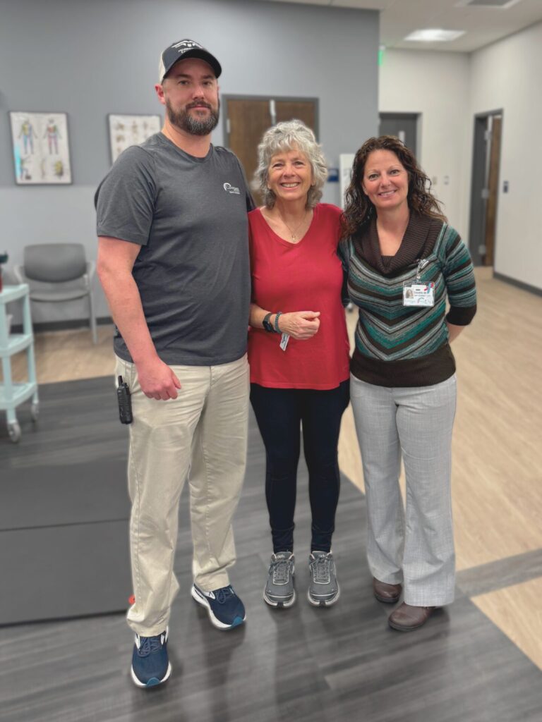 Three adults standing and smiling together in a medical or physical therapy office. The man on the left wears a gray T-shirt and cap, the woman in the middle wears a red top, and the woman on the right wears a brown sweater and slacks.