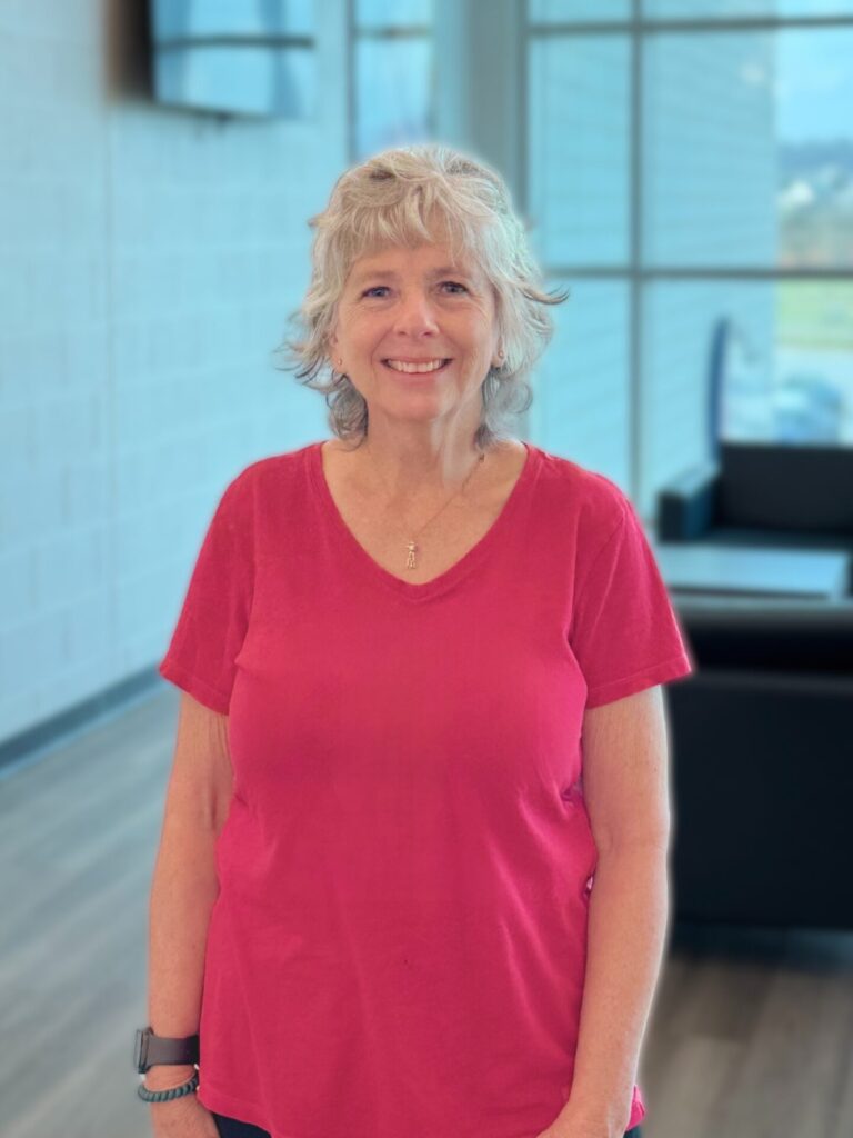 An older woman with curly gray hair and a red short-sleeve shirt stands smiling in a modern, well-lit room with large windows and black furniture in the background.