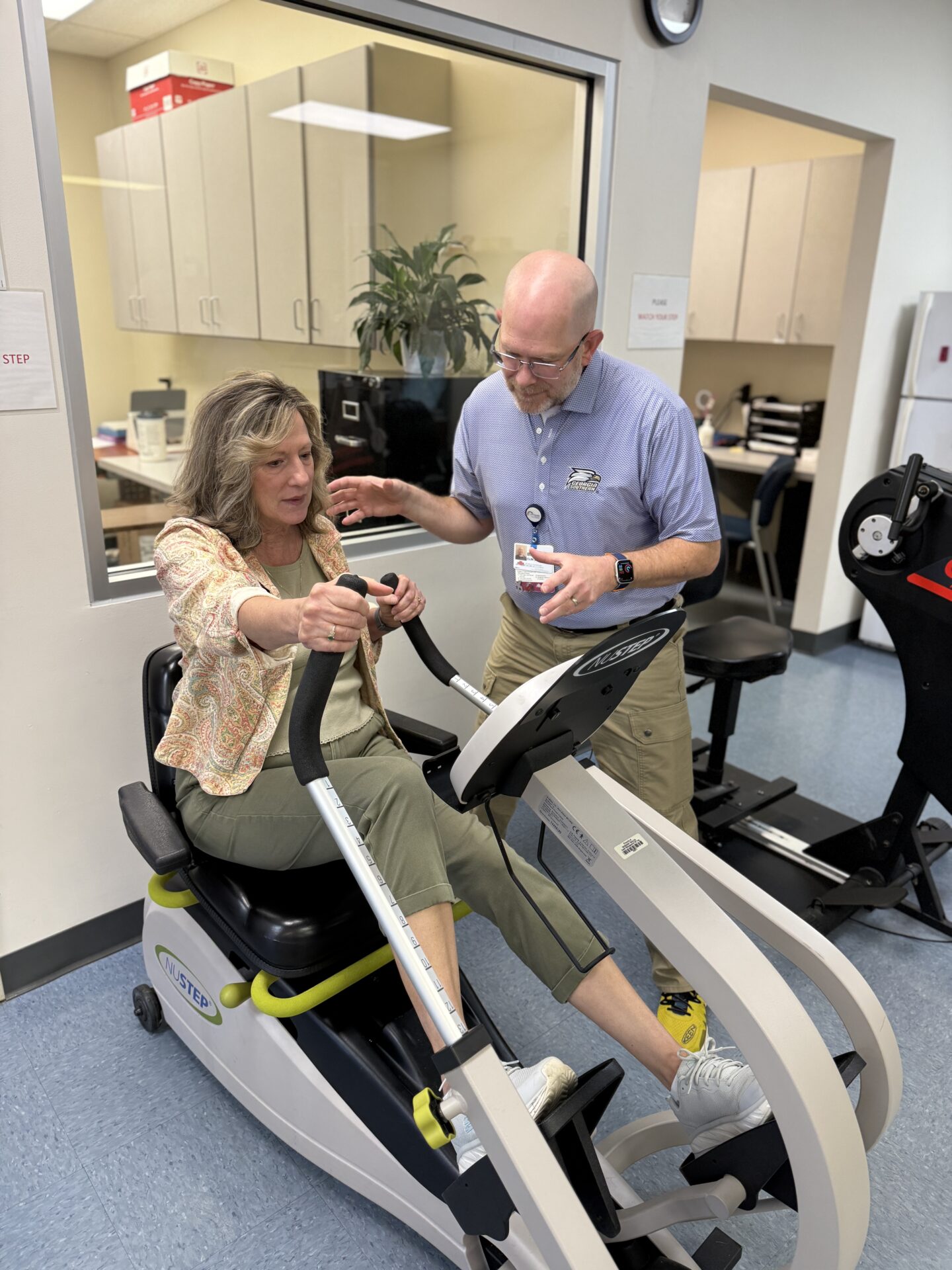 A woman exercises on a recumbent stepper machine while a male physical therapist stands beside her, offering guidance in a clinic setting.