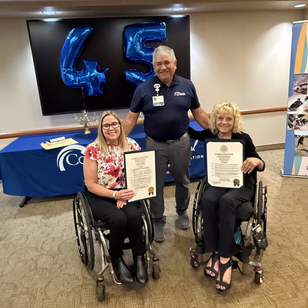 Two women in wheelchairs hold certificates while a man stands behind them, smiling. Behind them is a table with a blue cloth, a book, and large blue balloons shaped as the number 45.