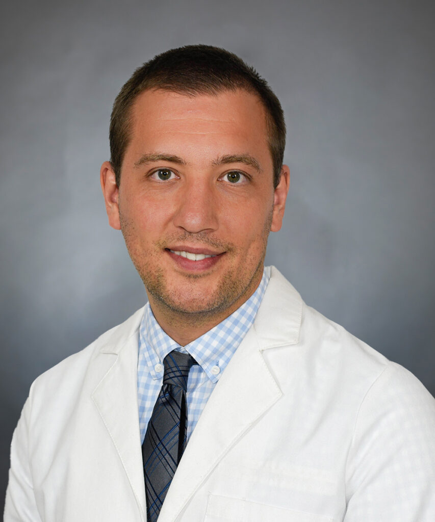 A man wearing a white lab coat, blue and white checkered shirt, and dark patterned tie, posing in front of a plain gray background.