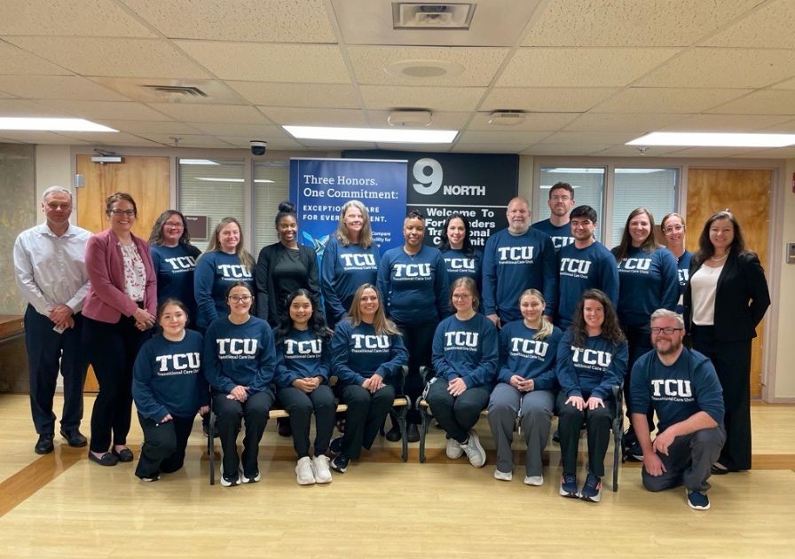 A group of twenty people, some standing and some seated, pose indoors. Many wear matching navy blue TCU shirts. They are in a hallway with wood floors and a sign behind them that reads 9 North.