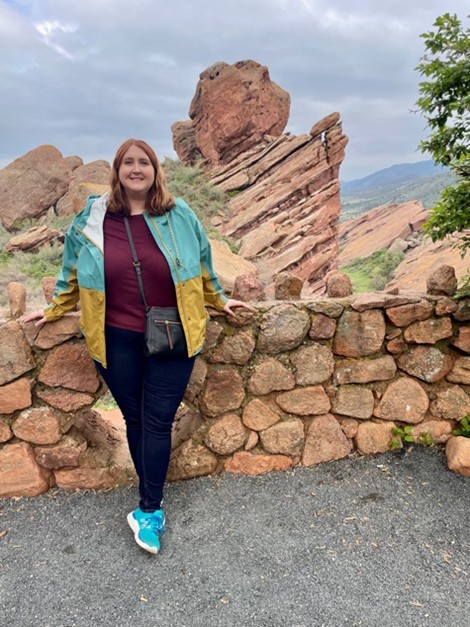 A woman in a colorful jacket and blue sneakers stands smiling in front of a stone wall, with rugged red rock formations and cloudy skies in the background.