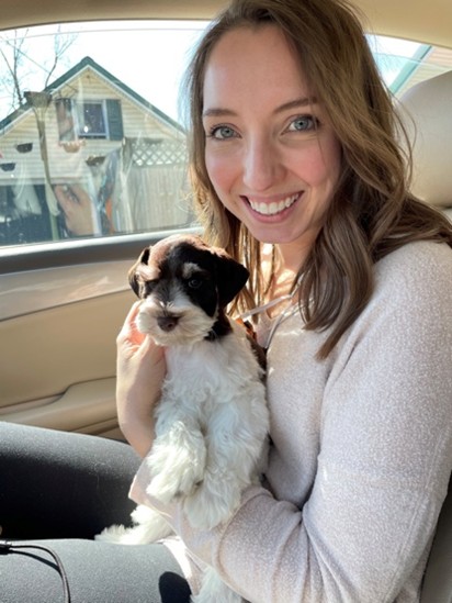 A smiling woman with light brown hair sits in a car holding a small black and white puppy on her lap. Sunlight shines through the window, and a house is visible outside.