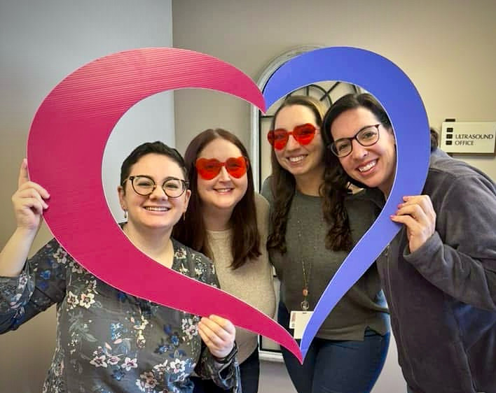 Four smiling women pose together indoors, holding a large pink and blue heart-shaped frame. Two are wearing red heart-shaped glasses. A sign in the background reads Ultrasound Office.