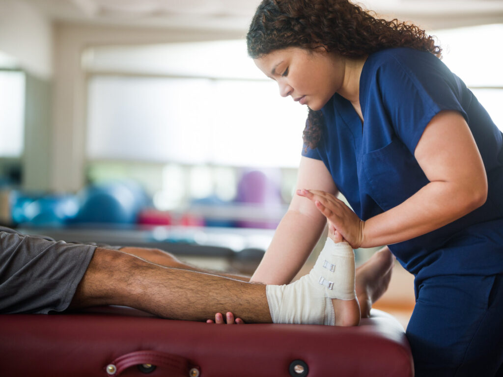 A physical therapist in blue scrubs gently stretches the ankle of a patient with a bandaged foot, who is lying on a therapy table in a bright, modern clinic.