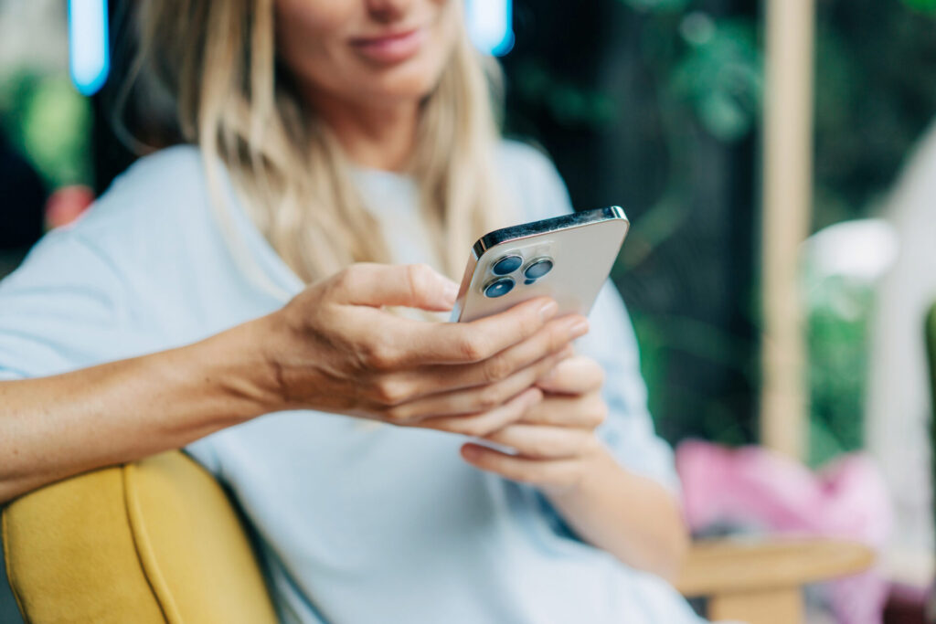 A woman with long blonde hair sits on a yellow chair, holding and using a smartphone. The image focuses on her hands and phone, with her face slightly blurred in the background.