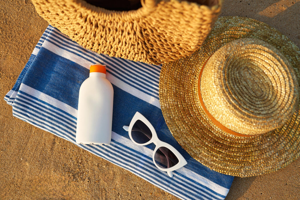 A woven straw hat, a matching bag, white sunglasses, and a bottle of sunscreen rest on a blue and white striped towel on sandy beach.