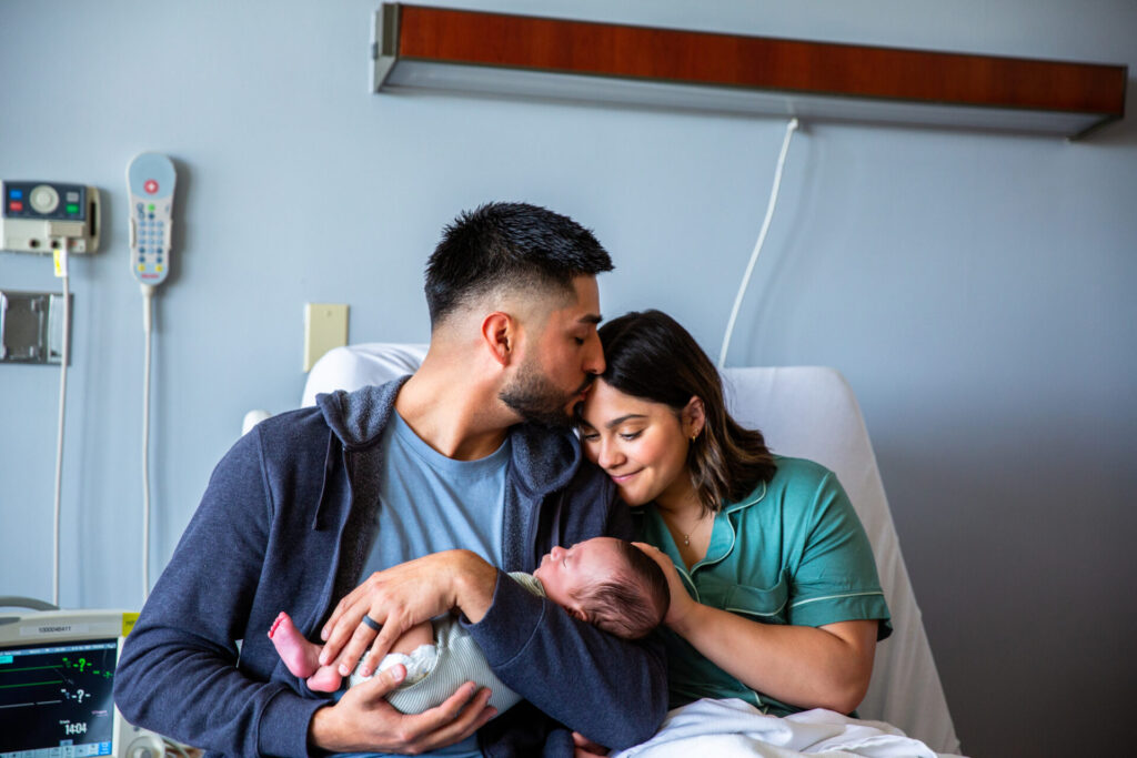 A man kisses a woman’s forehead as they both sit on a hospital bed, lovingly holding a newborn baby wrapped in a blanket. Medical equipment is visible in the background.