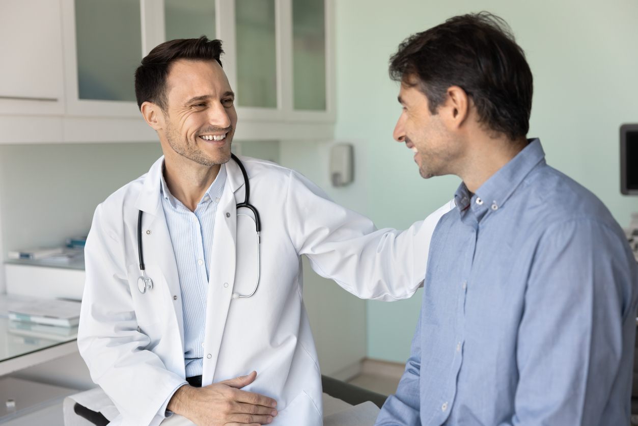 A doctor with a stethoscope smiles and reassures a patient by gently placing a hand on his shoulder during a consultation in a bright medical office.