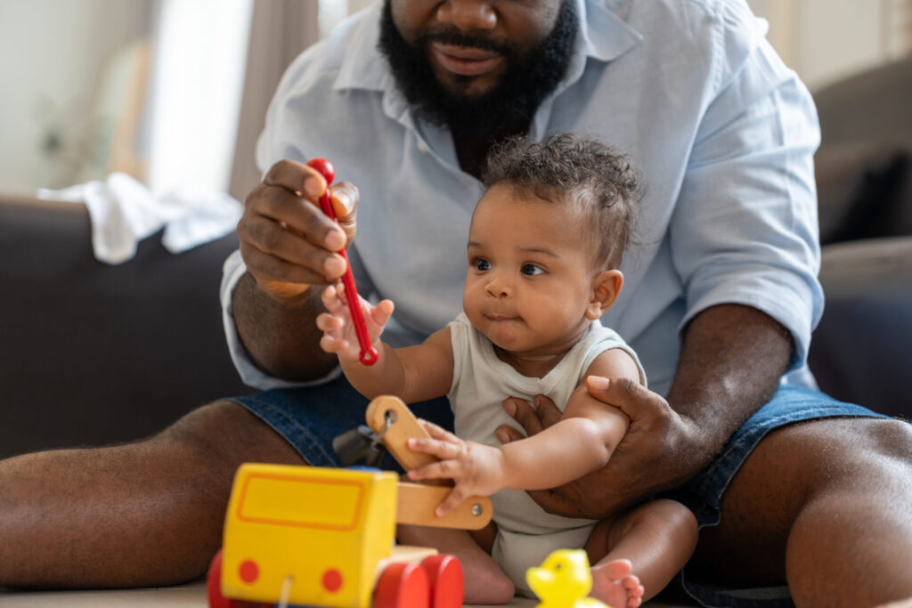 A man sits on the floor holding a baby, helping them play with a wooden toy truck and a red tool. The baby looks curious and engaged, surrounded by toys.