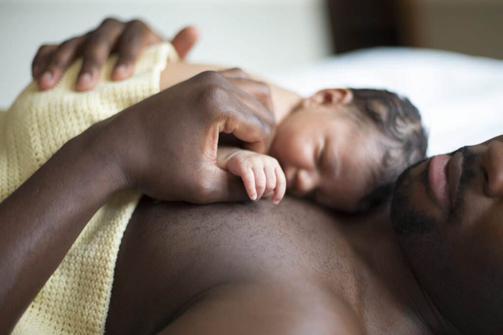 A baby sleeps peacefully on an adult’s bare chest, both partially covered with a yellow knitted blanket, capturing a tender moment of closeness and bonding.