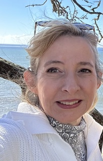 A woman with light hair, wearing a white sweater and patterned scarf, smiles outdoors by the sea beneath blue sky and branches—celebrating Michaela’s recovery from severe blockage with Covenant Health. Sunglasses rest on her head.