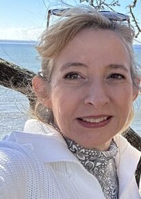 A woman with light hair and a scarf smiles at the camera outdoors, with the ocean and a blue sky in the background. She is wearing a white jacket and sunglasses are perched on her head.