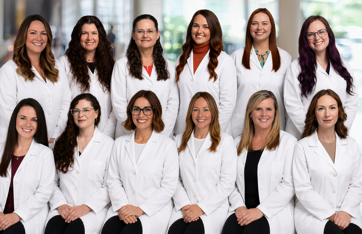 Twelve women in white lab coats pose together, smiling in two rows, against a blurred indoor background that suggests a professional or medical setting specializing in Midwife Services.
