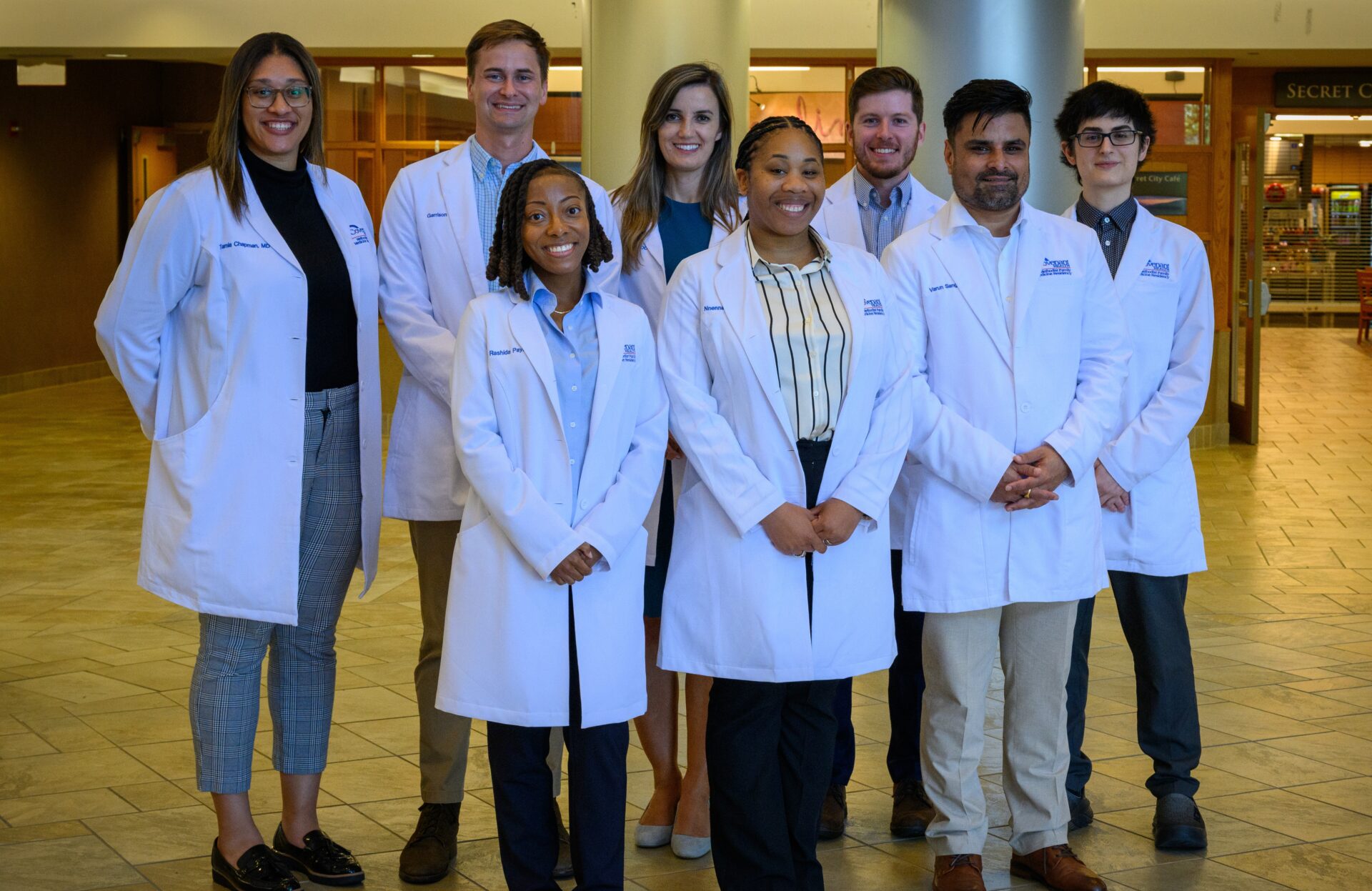 Eight people wearing white lab coats stand together indoors, smiling at the camera. The background shows a spacious, well-lit hallway with pillars and a sign reading SECRET.