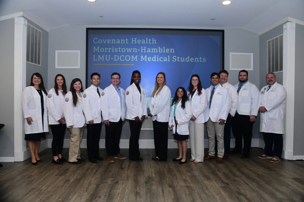 Thirteen people in white coats stand side by side in front of a screen that reads Covenant Health Morristown-Hamblen LMU-DCOM Medical Students in a well-lit room with light-colored walls and a wood floor.