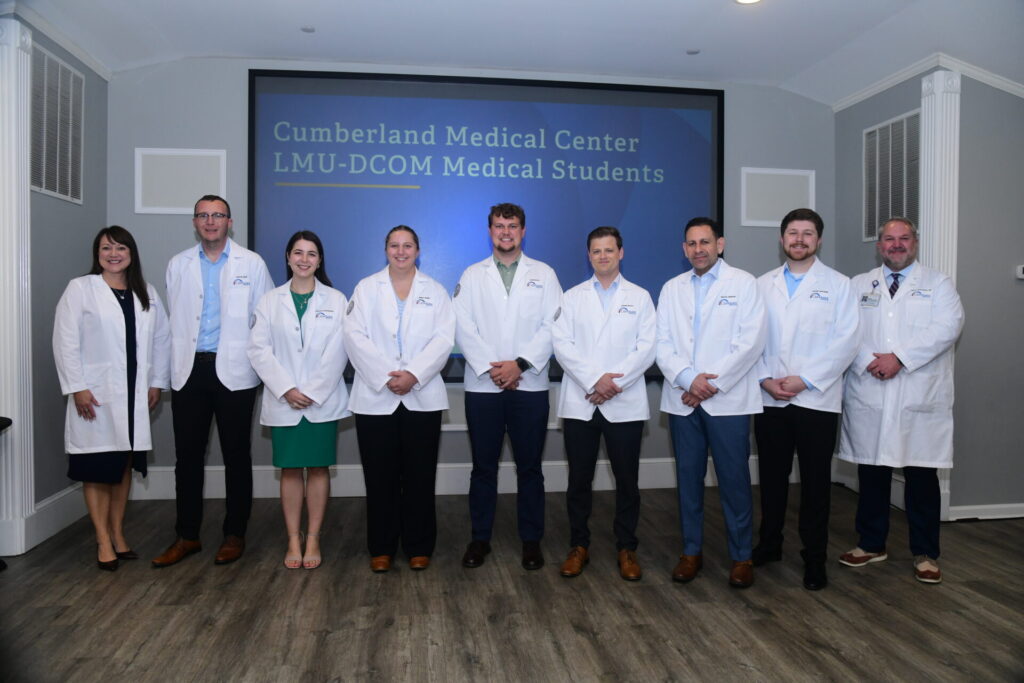 Nine people in white coats stand in front of a screen that reads Cumberland Medical Center LMU-DCOM Medical Students. They are lined up in a conference room, smiling at the camera.