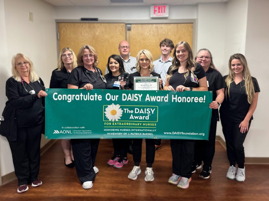 Ten people, mostly in black scrubs, stand in a hallway holding a green banner that reads Congratulate Our DAISY Award Honoree! One person in the center holds a certificate. They are smiling and facing the camera.