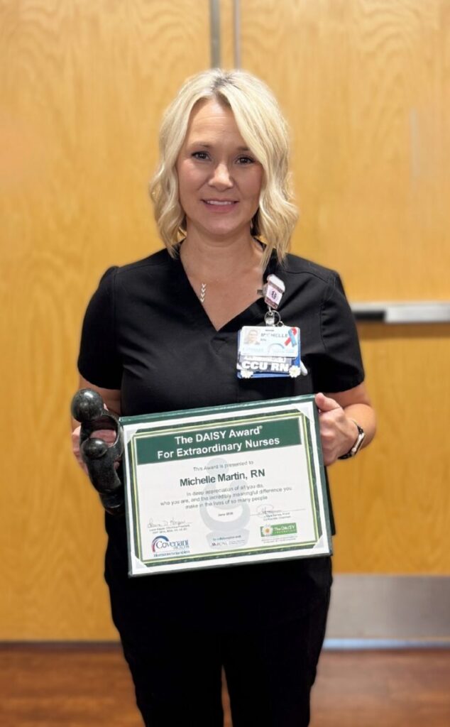 A woman in black scrubs stands indoors holding a framed certificate and a small award, smiling at the camera. She is in front of closed double doors on a brown floor.