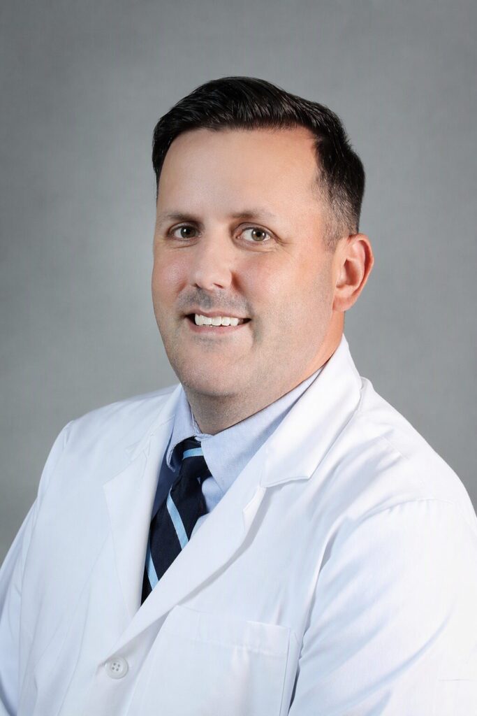 A man wearing a white lab coat, dress shirt, and striped tie smiles at the camera against a plain gray background.