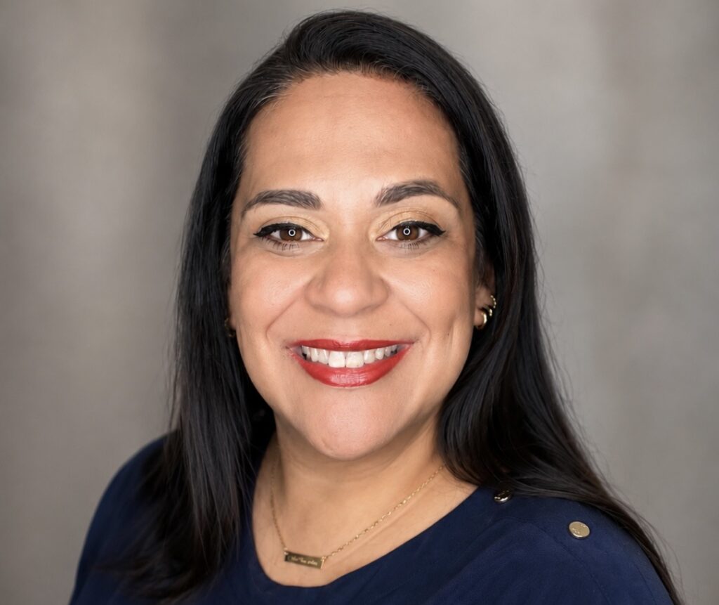A woman with long dark hair smiles at the camera. She is wearing a navy blue top with gold buttons on the shoulder, a gold necklace, and red lipstick. The softly blurred gray background suggests a professional setting, perhaps taken at the metabolic clinic in Oak Ridge.