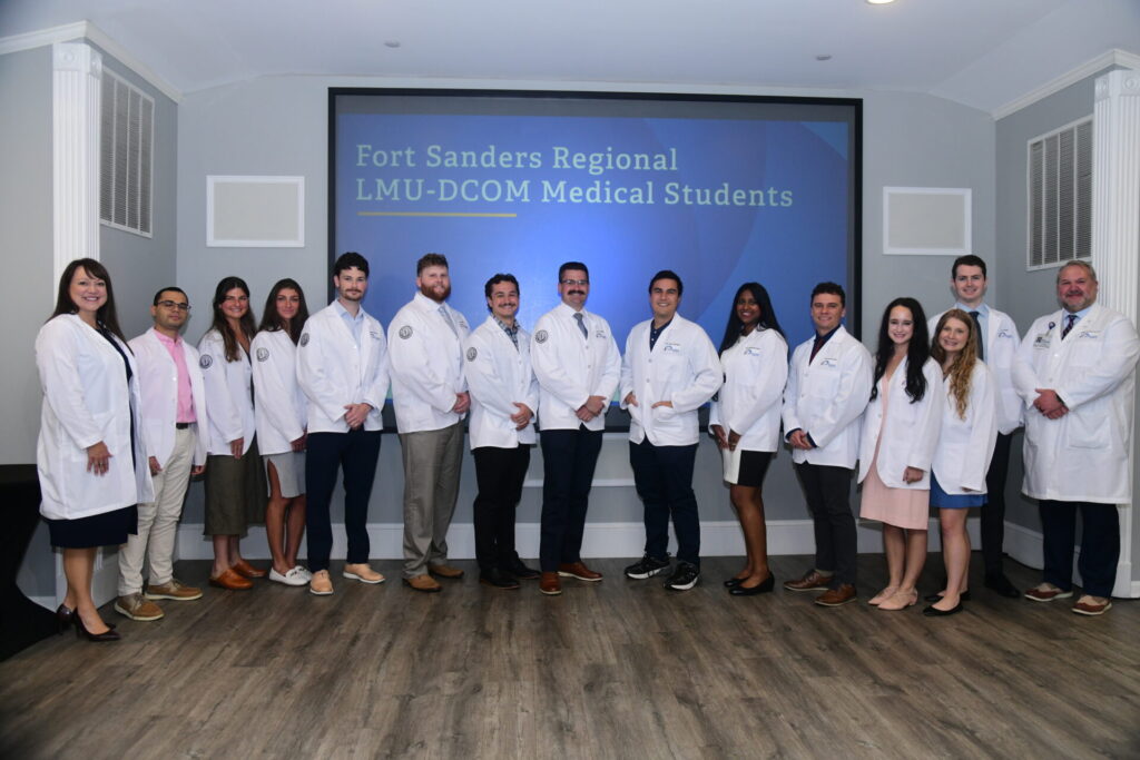 A group of medical students and faculty in white coats stand in a line indoors, smiling at the camera. A screen behind them reads Fort Sanders Regional LMU-DCOM Medical Students.