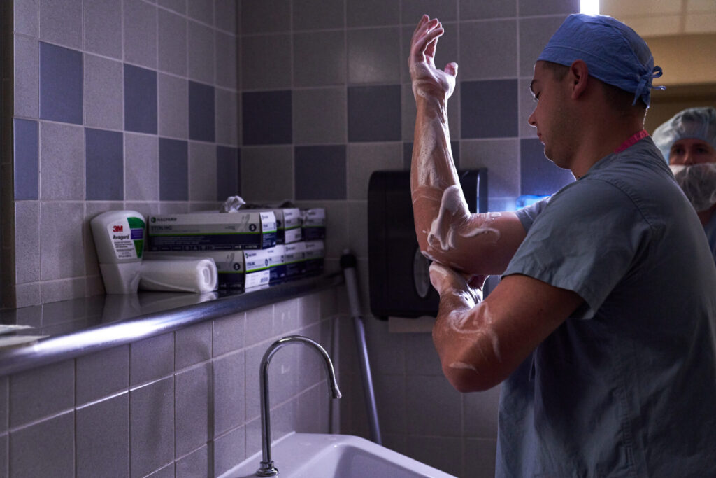 A medical professional in scrubs and a cap scrubs his arms with soap at a sink in a sterile hospital environment, likely preparing for surgery. Medical supplies are visible on a shelf in the background.