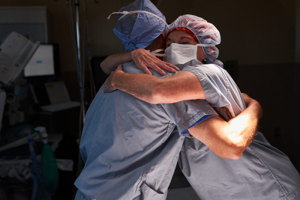 Two healthcare workers in scrubs, surgical caps, and masks embrace each other in a hospital setting, with medical equipment visible in the background.