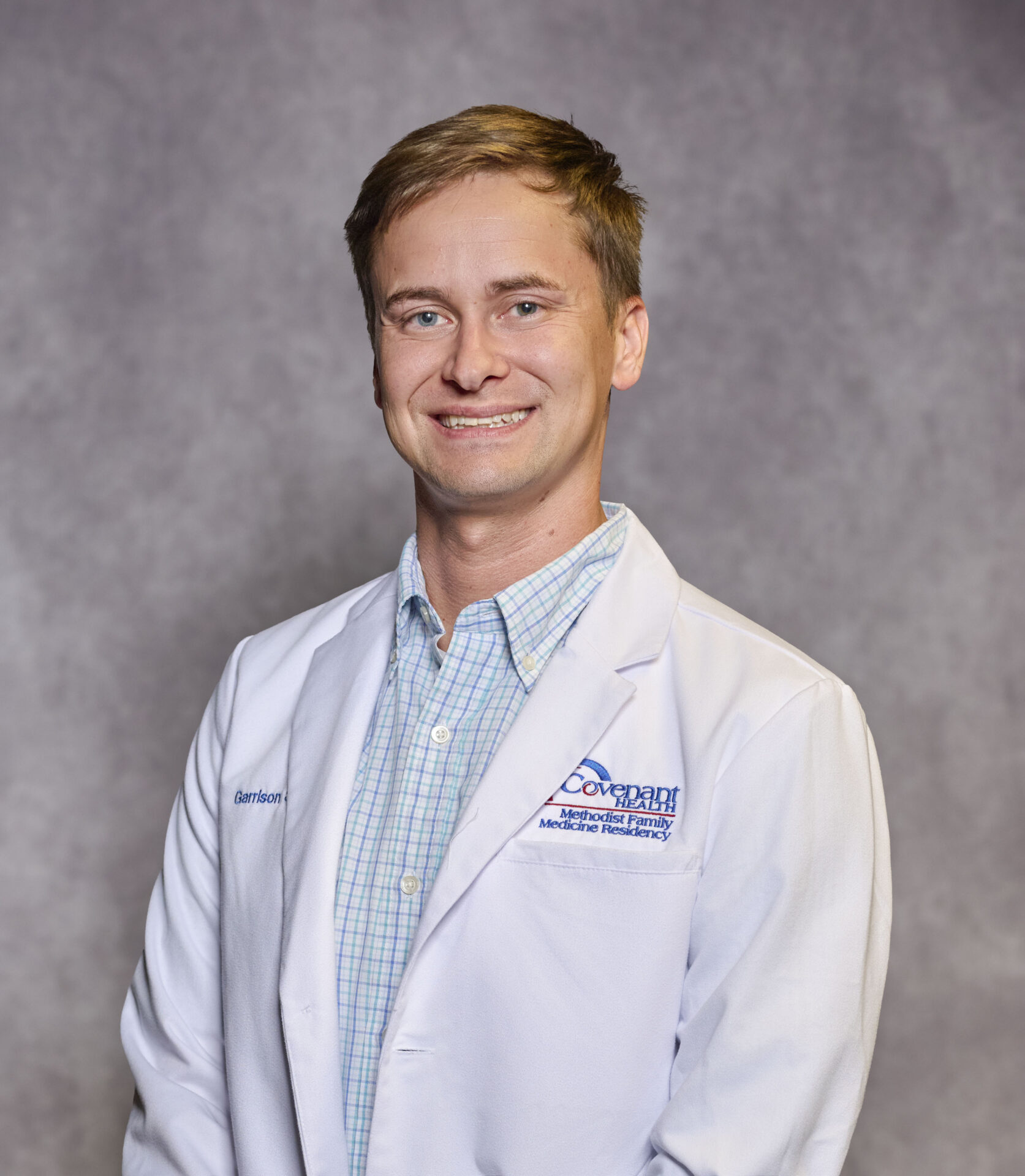 A smiling man in a white medical coat stands against a plain gray background. The coat has blue embroidery and a name tag on it. He is wearing a light blue plaid shirt underneath.