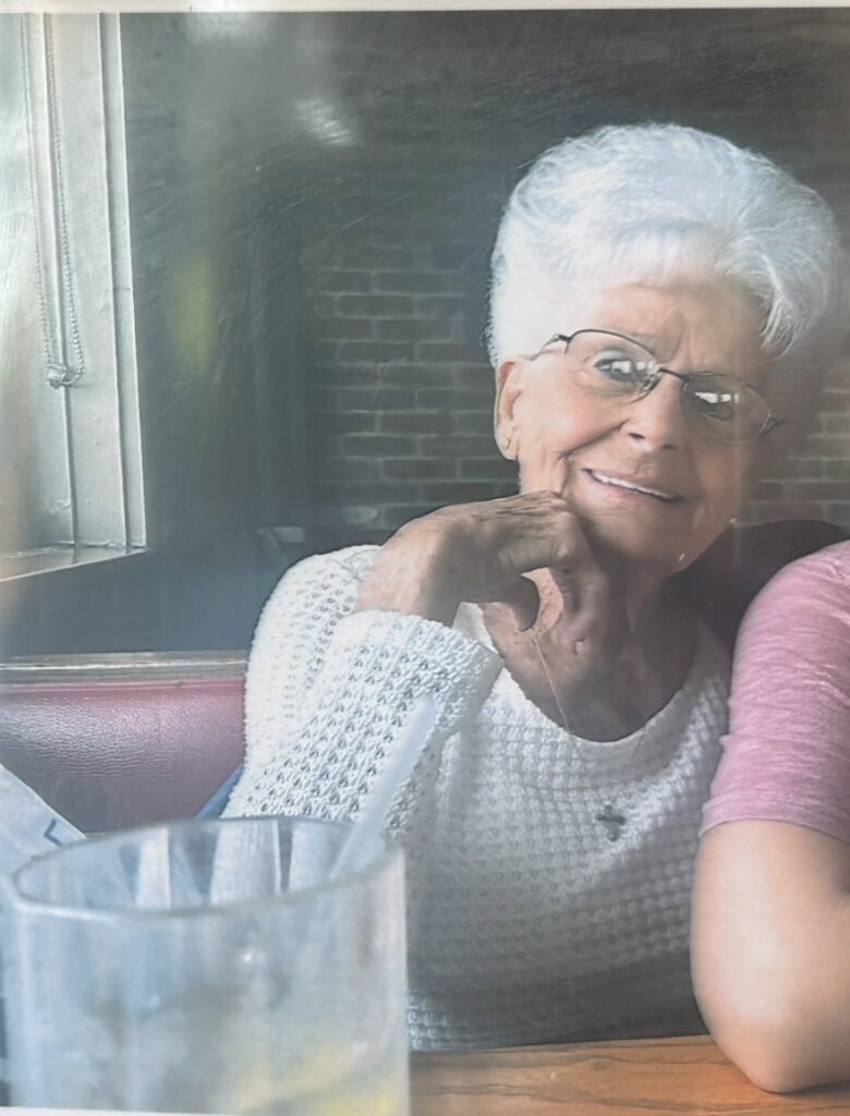 An elderly woman with white hair and glasses smiles while sitting at a restaurant table next to someone. She wears a hip white sweater, and there&rsquo;s a glass of ice water with a straw on the table in front of her.