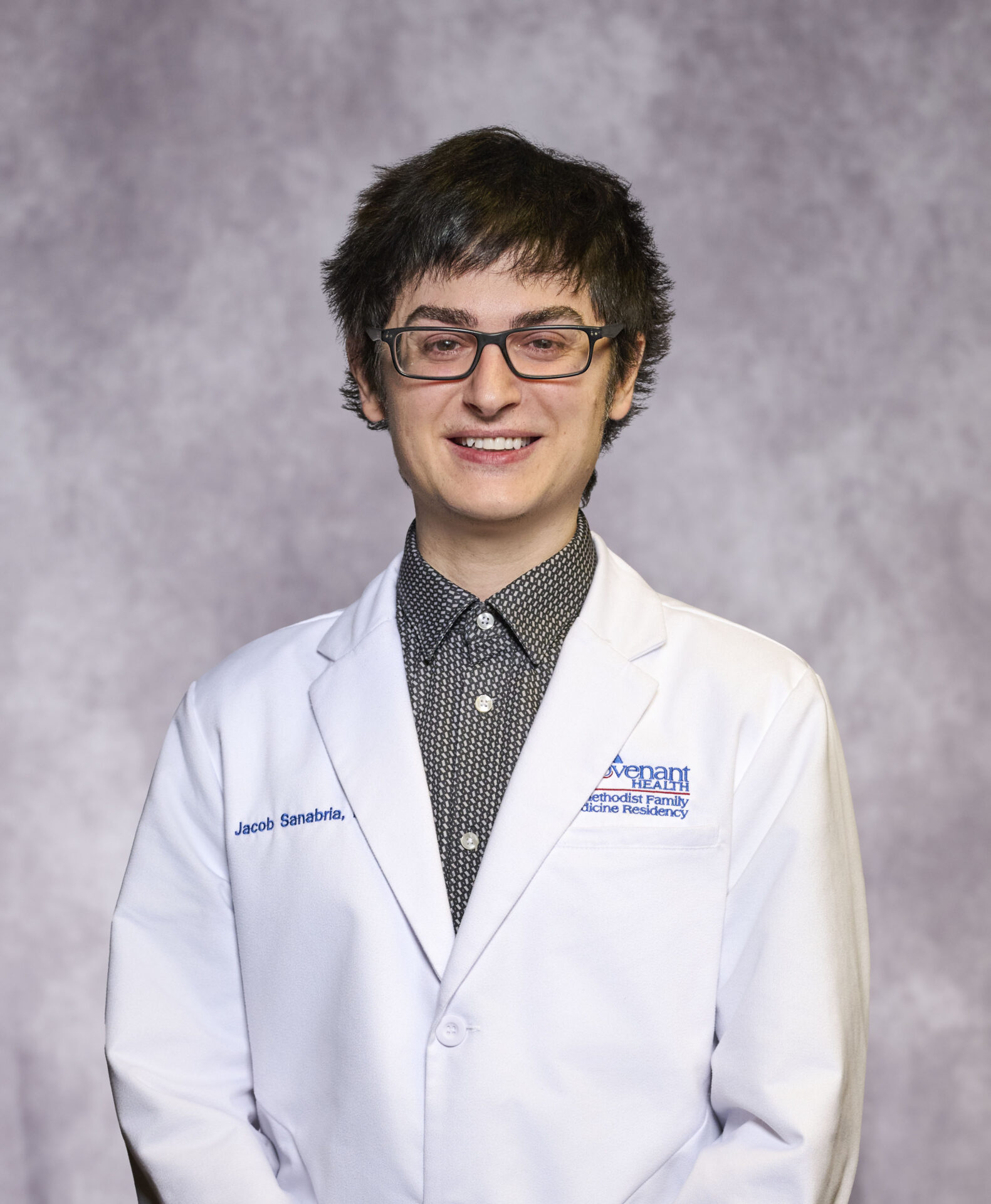 A person with short, dark hair and glasses is smiling, wearing a white lab coat over a patterned shirt, standing in front of a light gray textured background.