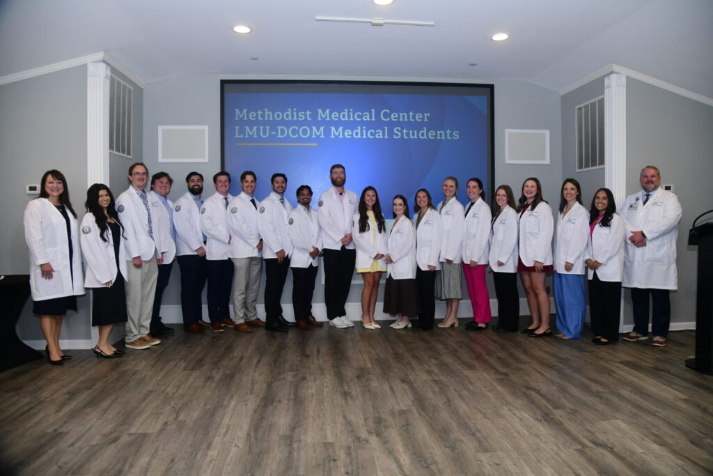 A group of medical students and faculty in white coats stand in a line for a photo in front of a screen that reads Methodist Medical Center LMU-DCOM Medical Students.