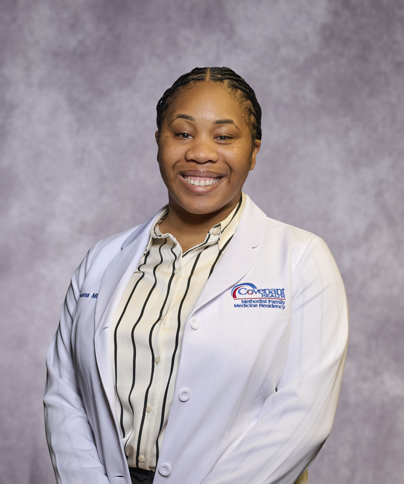 A woman in a white medical coat with a name badge smiles in front of a light purple background. She is wearing a striped blouse and has her hair braided back.