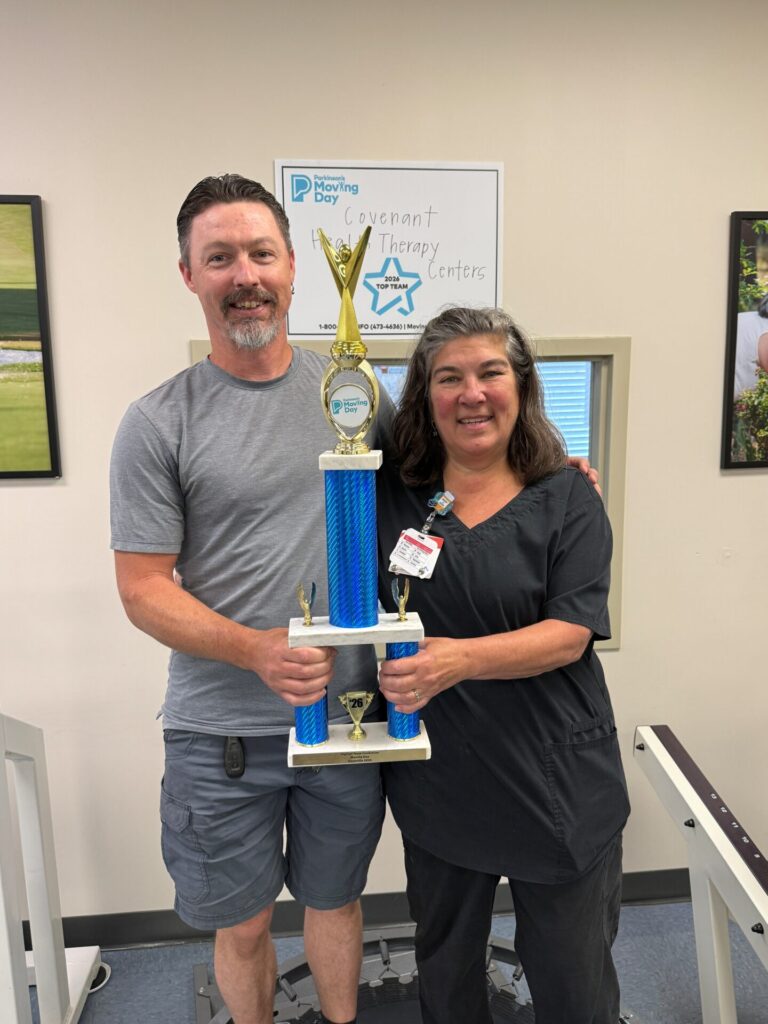Two people stand indoors smiling and holding a large blue and gold trophy. Behind them is a sign reading Covenant Therapy Centers. They appear proud and happy, likely celebrating a recent achievement.