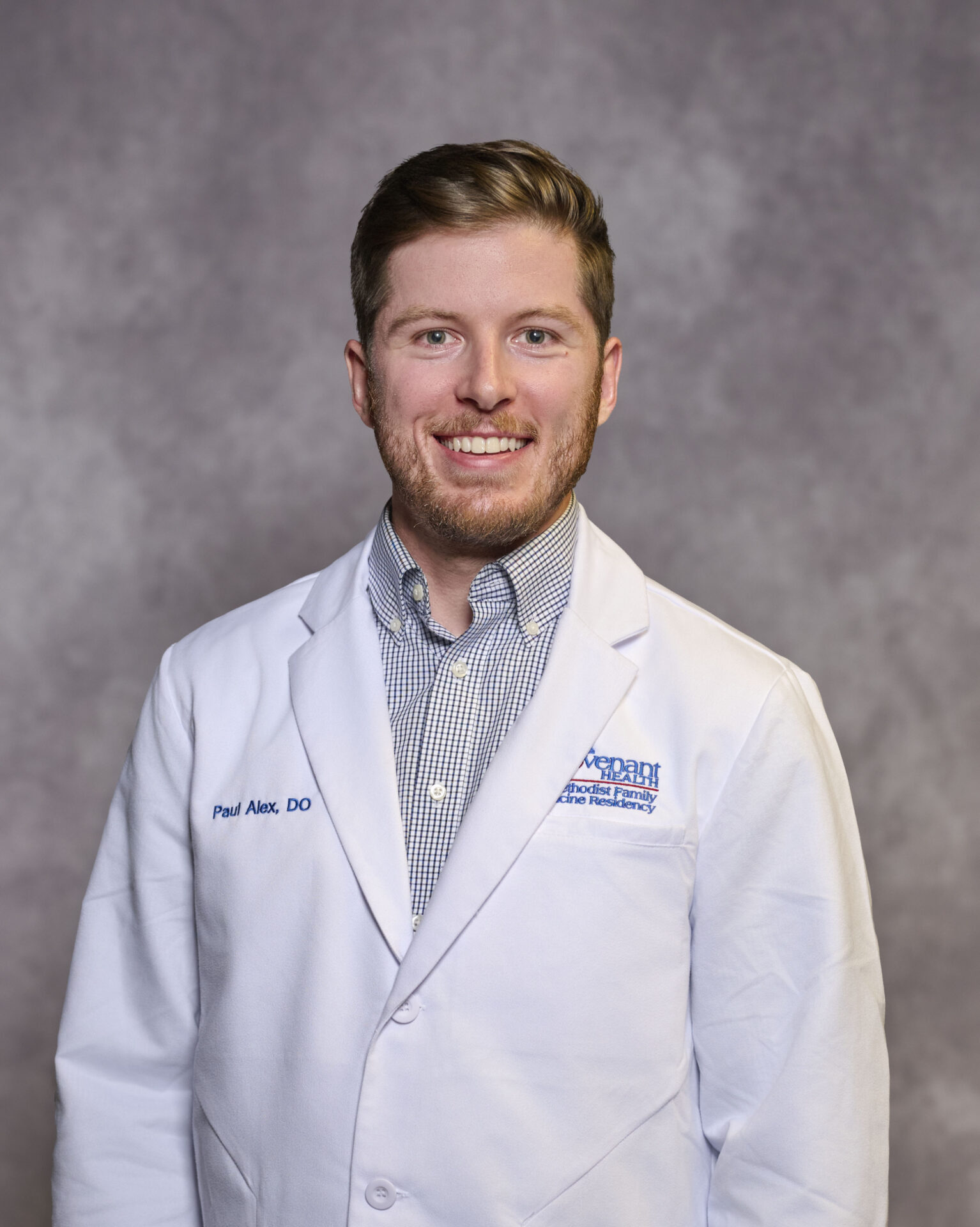 A man with short light brown hair and a beard smiles, wearing a white doctor’s coat over a checkered shirt, standing in front of a gray mottled background.