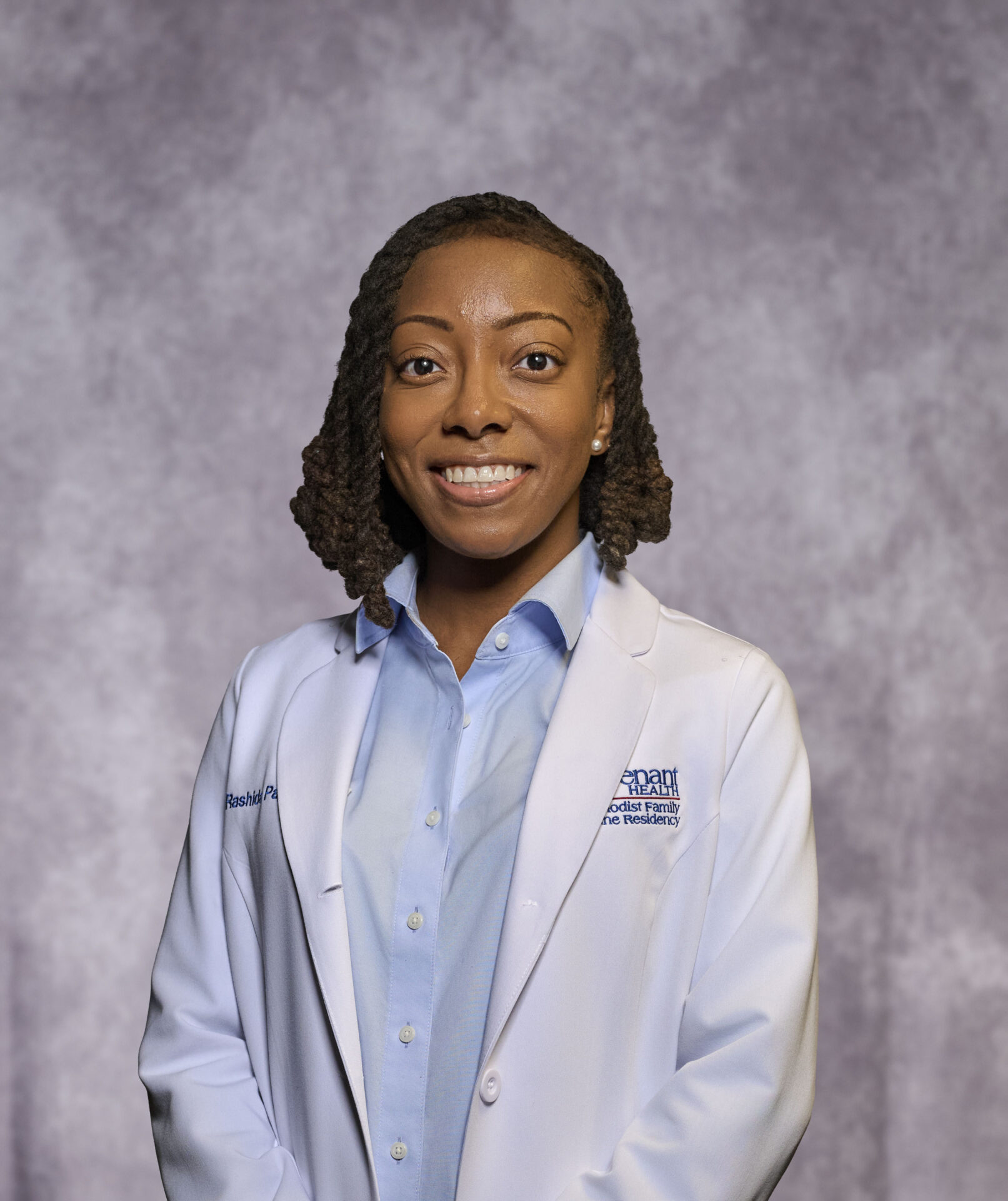 A woman wearing a white medical coat and blue shirt stands smiling in front of a light gray textured backdrop.