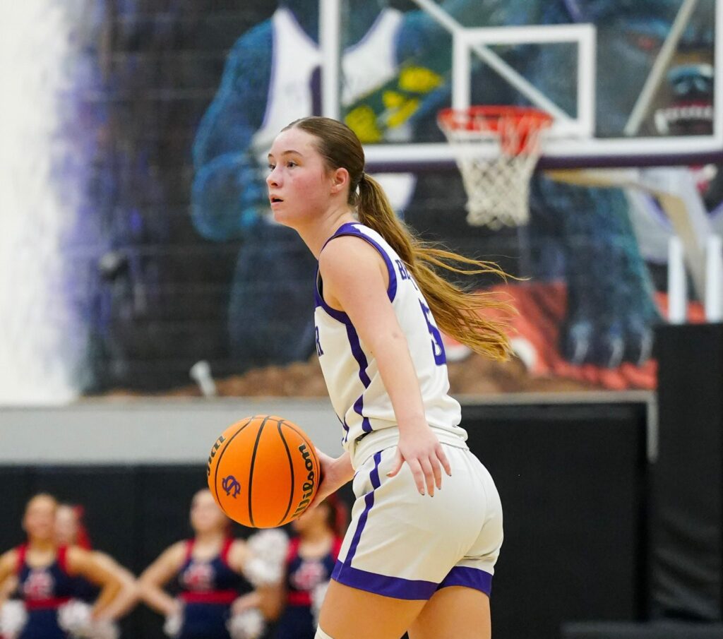 A female basketball player in a white and purple uniform dribbles a basketball on a court, with a colorful mural and blurred spectators in the background.
