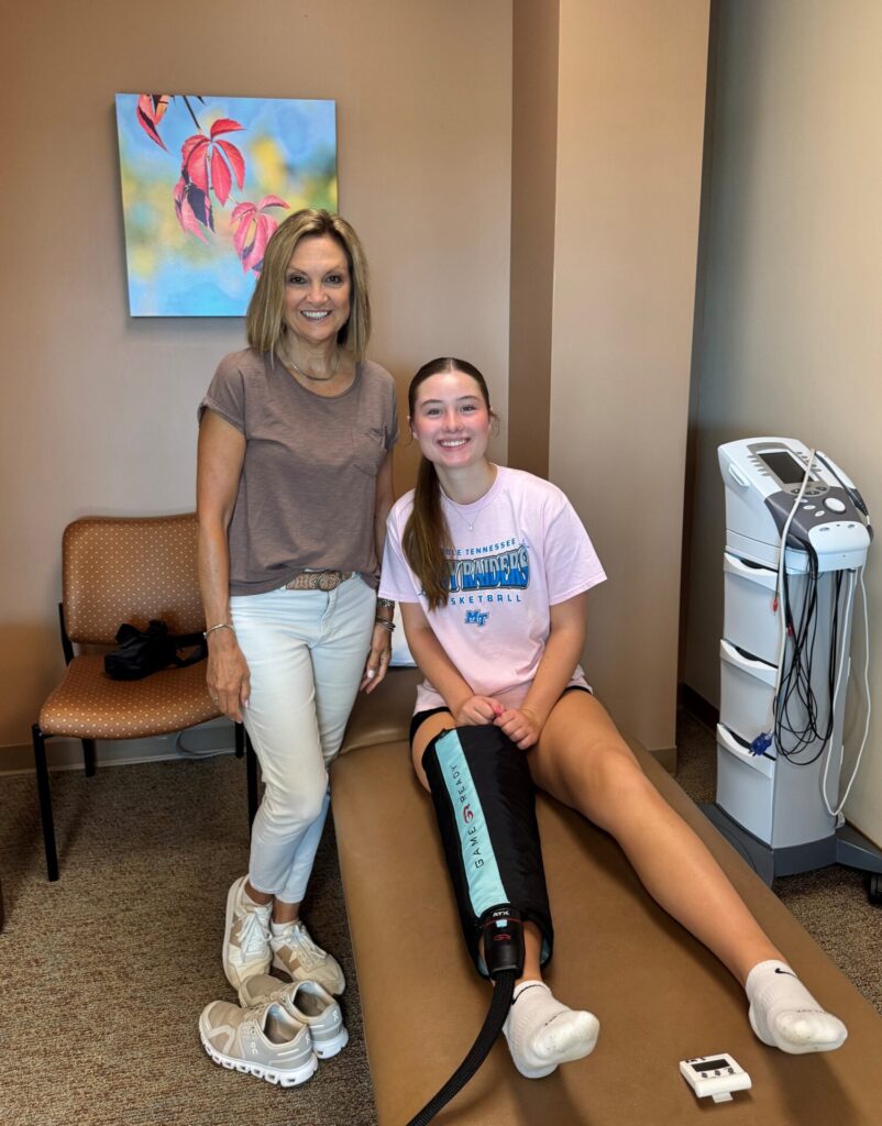 Two women in a medical office; one is seated on an exam table with her leg in a brace, smiling. The other stands beside her, also smiling. Medical equipment and artwork are visible in the background.