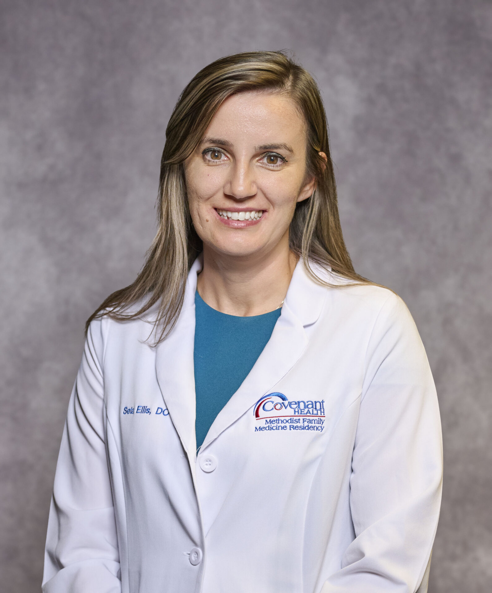 A woman with long light brown hair wearing a white medical coat over a blue top, standing in front of a gray background and smiling at the camera.